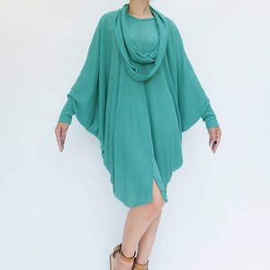 K&ouml;nnte beinhalten: Ein t&uuml;rkisfarbenes, drapiertes Kleid mit einem Wasserfallausschnitt und langen &Auml;rmeln. Das Kleid hat einen asymmetrischen Saum und wird mit braunen Sandalen getragen. Das Model tr&auml;gt eine gro&szlig;e, dunkle Sonnenbrille.