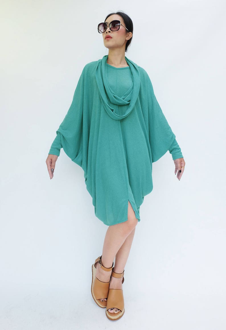 K&ouml;nnte beinhalten: Ein t&uuml;rkisfarbenes, drapiertes Kleid mit einem Wasserfallausschnitt und langen &Auml;rmeln. Das Kleid hat einen asymmetrischen Saum und wird mit braunen Sandalen getragen. Das Model tr&auml;gt eine gro&szlig;e, dunkle Sonnenbrille.