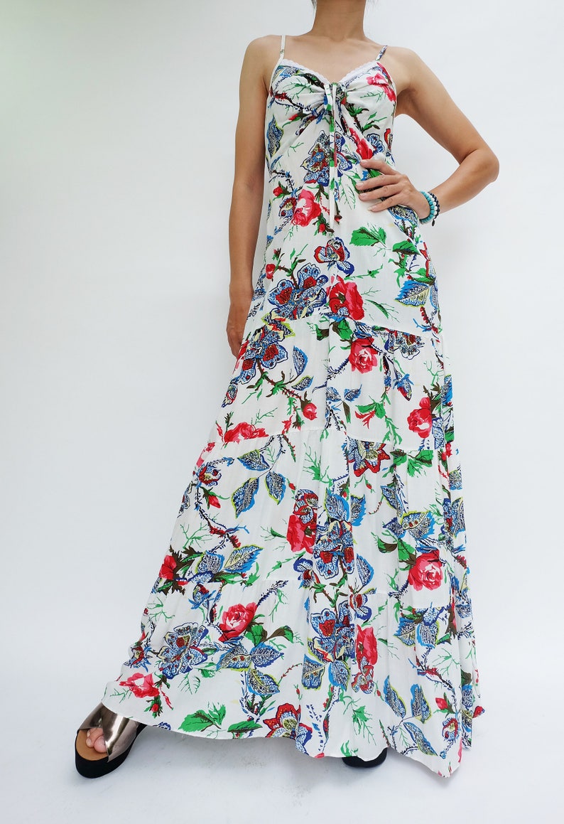 May include: White maxi dress with a floral pattern featuring red roses, blue paisley, and green leaves. The dress has spaghetti straps and a tiered skirt. The top has a tie detail at the bust.