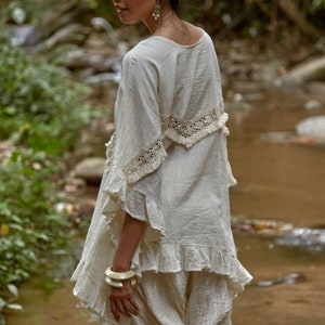 May include: A woman wearing a white, crocheted top with a ruffled hem and white pants. She is standing in a forest setting near a stream.