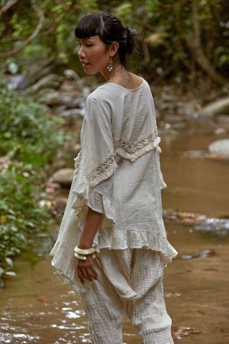 May include: A woman wearing a white, crocheted top with a ruffled hem and white pants. She is standing in a forest setting near a stream.