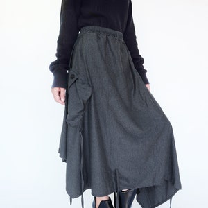 May include: A dark gray long skirt with a large cargo pocket on the left side. The skirt has a drawstring waist and a slightly asymmetrical hemline. The skirt is paired with black ankle boots.