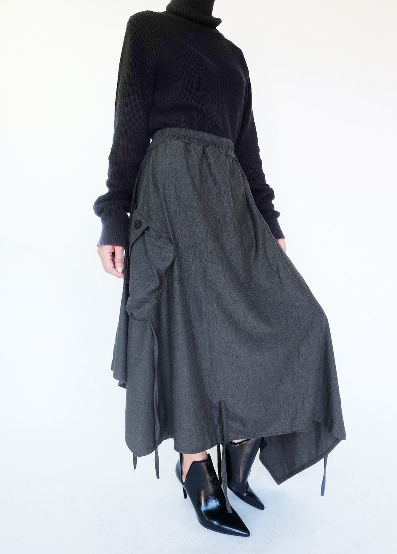 May include: A dark gray long skirt with a large cargo pocket on the left side. The skirt has a drawstring waist and a slightly asymmetrical hemline. The skirt is paired with black ankle boots.