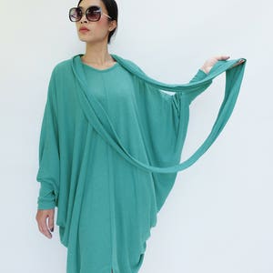 K&ouml;nnte beinhalten: Ein t&uuml;rkisfarbenes, lang&auml;rmeliges Kleid mit drapiertem Ausschnitt und flie&szlig;endem, asymmetrischem Design. Das Kleid hat eine lockere, bequeme Passform und einen Schlitz an der Seite. Das Model tr&auml;gt eine gro&szlig;e, dunkle Sonnenbrille.