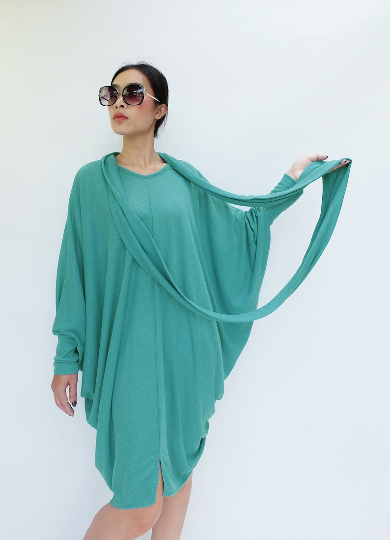K&ouml;nnte beinhalten: Ein t&uuml;rkisfarbenes, lang&auml;rmeliges Kleid mit drapiertem Ausschnitt und flie&szlig;endem, asymmetrischem Design. Das Kleid hat eine lockere, bequeme Passform und einen Schlitz an der Seite. Das Model tr&auml;gt eine gro&szlig;e, dunkle Sonnenbrille.
