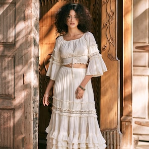 Puede incluir: Una falda maxi blanca de capas con un delicado ribete de encaje y un top corto blanco a juego con un lazo en la cintura. La modelo est&aacute; de pie frente a una puerta de madera.