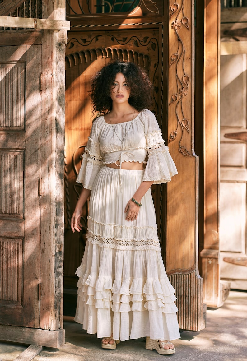 Puede incluir: Una falda maxi blanca de capas con un delicado ribete de encaje y un top corto blanco a juego con un lazo en la cintura. La modelo est&aacute; de pie frente a una puerta de madera.