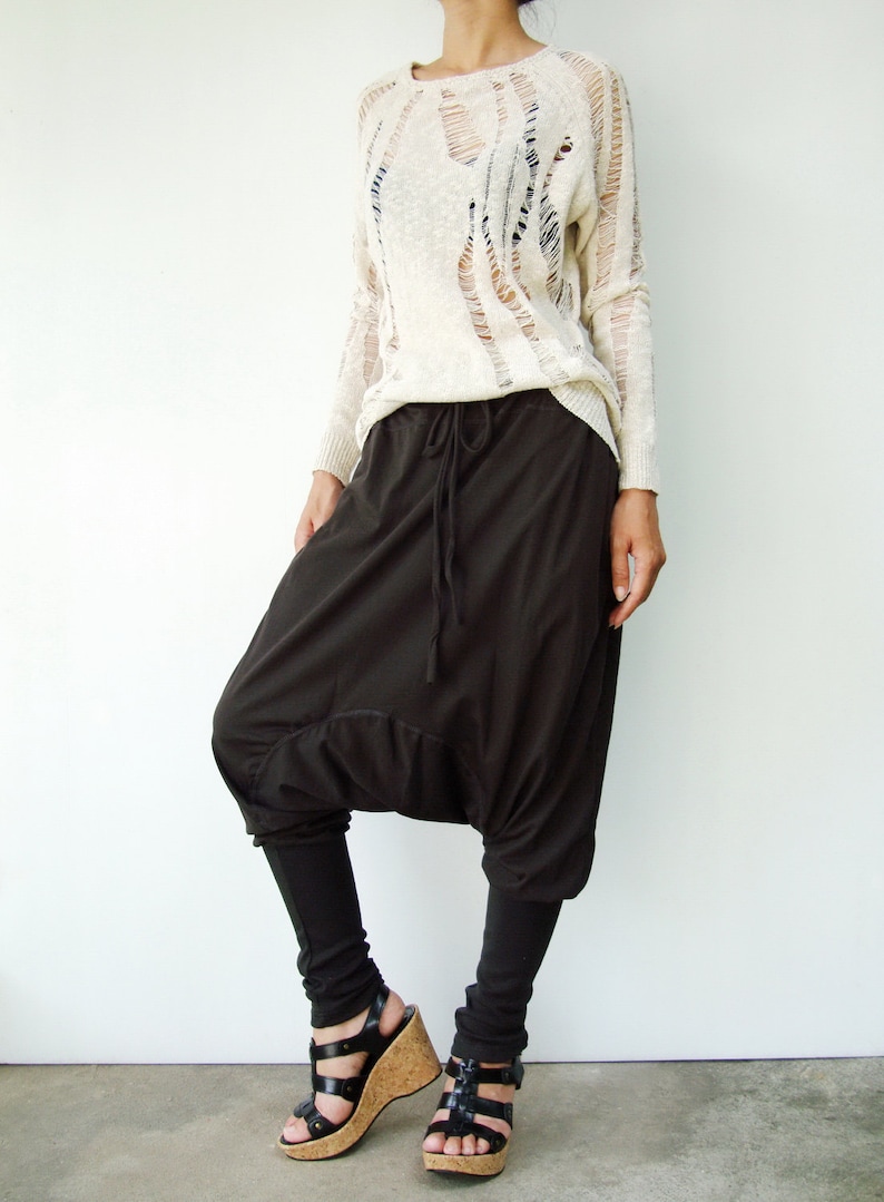 May include: A woman is wearing a white, long-sleeved, distressed sweater with a pair of brown, loose-fitting pants. She is wearing black sandals with cork wedges.