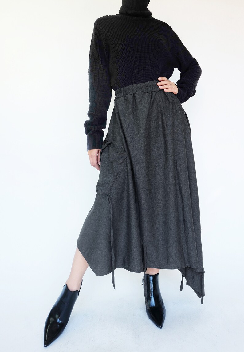 May include: A gray, asymmetrical skirt with a drawstring waist and a large cargo pocket on the left side. The skirt is paired with a black turtleneck sweater and black ankle boots.