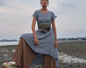 Könnte beinhalten: Eine Frau trägt ein hellblaues, locker sitzendes Kleid mit einem braunen Stoffgürtel, der zu einer Schleife gebunden ist. Sie trägt darunter eine braune, weite Hose und goldene Plateausandalen. Sie steht an einem felsigen Strand mit einem bewölkten Himmel im Hintergrund.