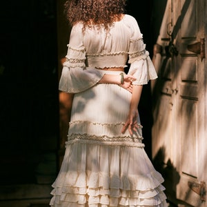 Puede incluir: Una falda maxi blanca de capas con un delicado ribete de encaje y un top corto blanco a juego con detalles de encaje. La falda tiene un estilo fluido y bohemio.