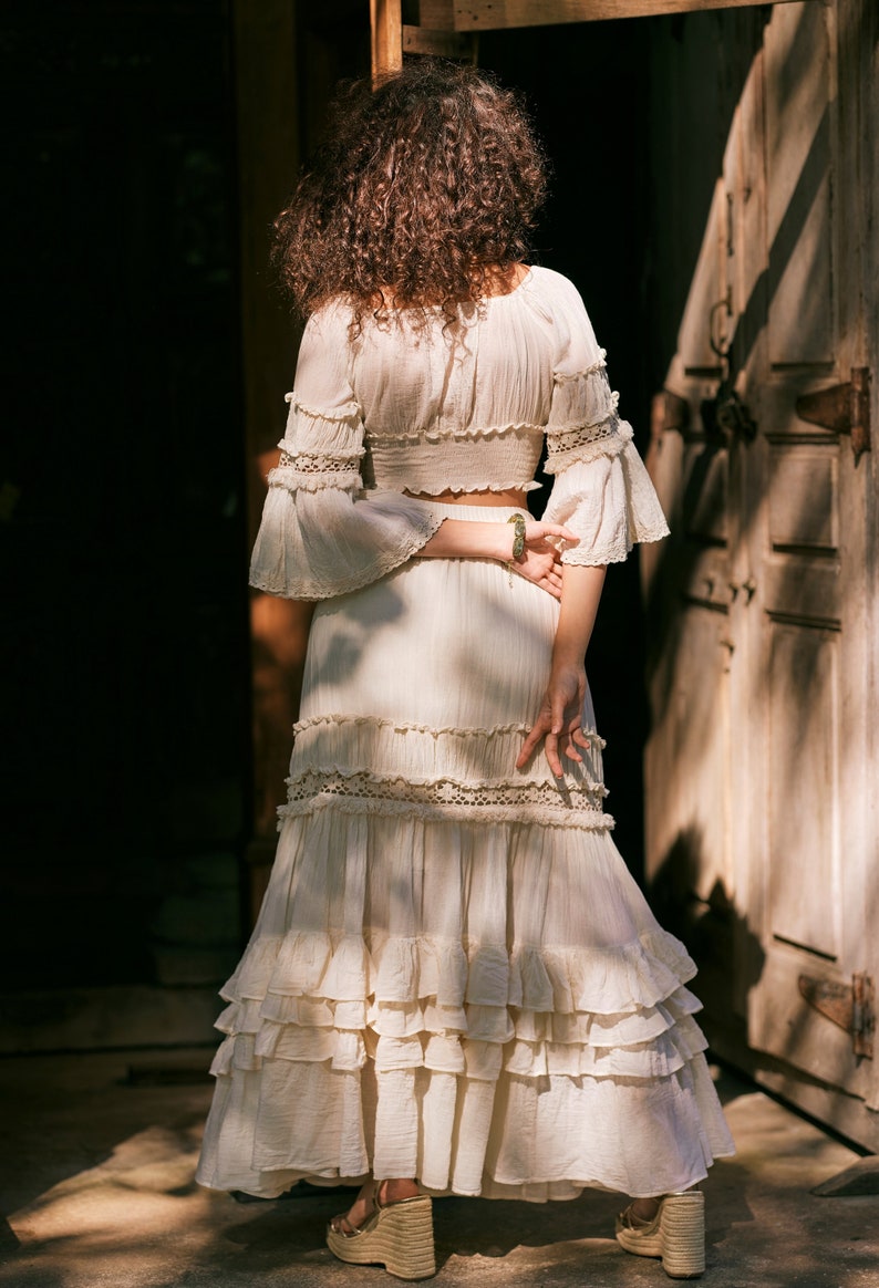 Puede incluir: Una falda maxi blanca de capas con un delicado ribete de encaje y un top corto blanco a juego con detalles de encaje. La falda tiene un estilo fluido y bohemio.