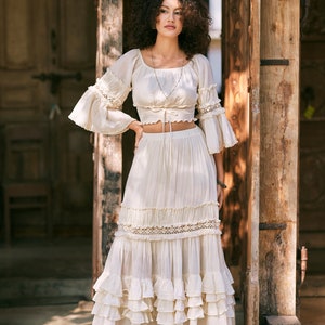 Puede incluir: Una falda maxi blanca de capas con ribete de encaje y un top corto blanco a juego con mangas de encaje. La modelo lleva el atuendo y est&aacute; de pie frente a una puerta de madera.