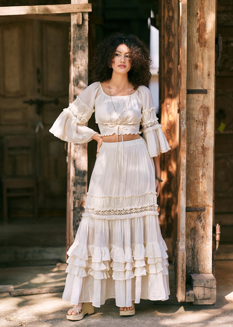 Puede incluir: Una falda maxi blanca de capas con ribete de encaje y un top corto blanco a juego con mangas de encaje. La modelo lleva el atuendo y est&aacute; de pie frente a una puerta de madera.