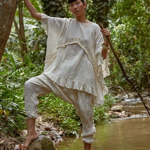 May include: A woman wearing a white, crocheted top and pants stands in a stream in a forest. She is holding a wooden stick in her right hand.