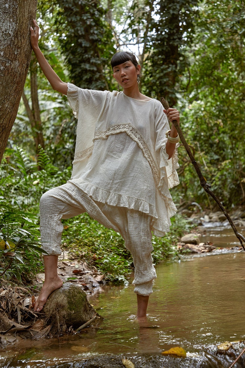 May include: A woman wearing a white, crocheted top and pants stands in a stream in a forest. She is holding a wooden stick in her right hand.