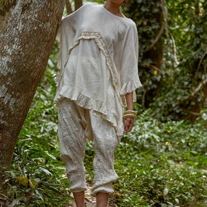 May include: A woman wearing a white, crocheted top with a ruffled hem and matching pants stands in a forest setting. She is wearing wooden bracelets on her wrist and is standing near a small stream.