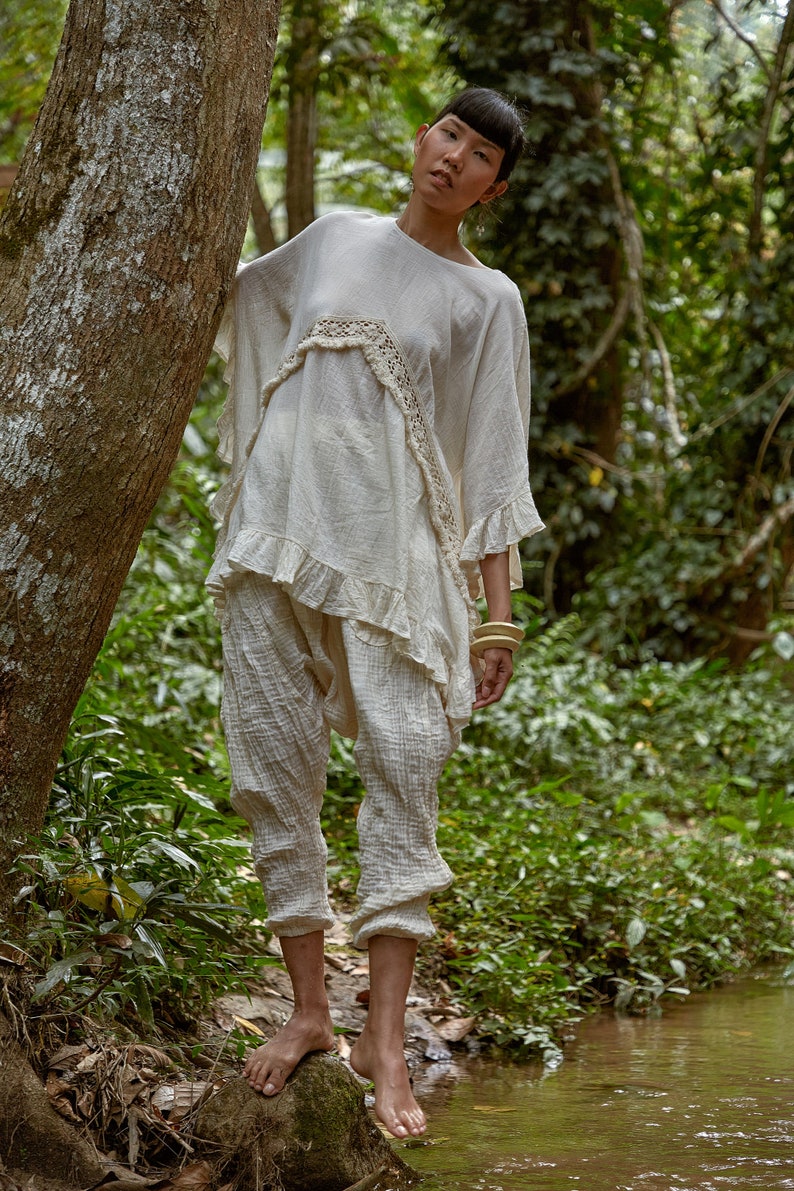 May include: A woman wearing a white, crocheted top with a ruffled hem and matching pants stands in a forest setting. She is wearing wooden bracelets on her wrist and is standing near a small stream.