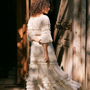Puede incluir: Una mujer lleva una falda maxi de crochet blanco con m&uacute;ltiples capas de volantes y ribetes de encaje. La falda es fluida y tiene un estilo bohemio.