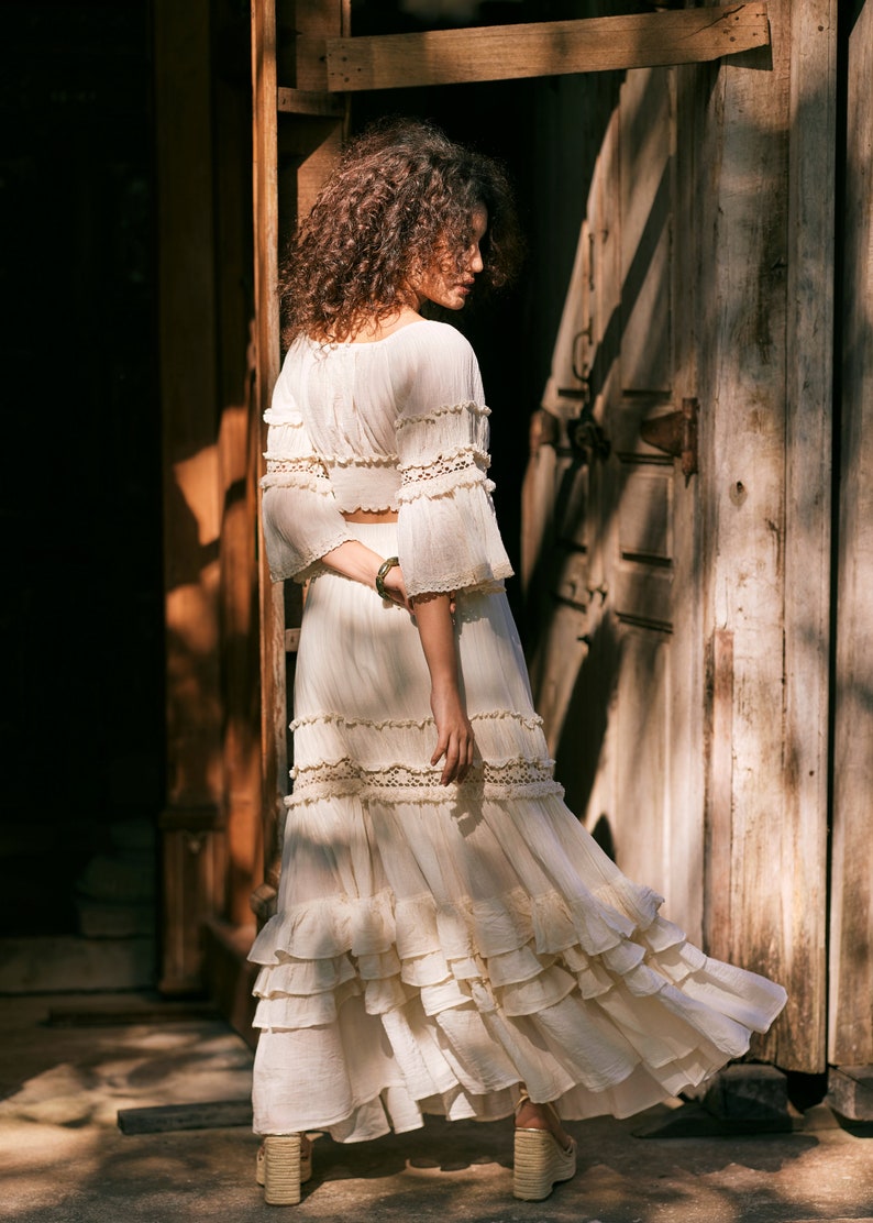 Puede incluir: Una mujer lleva una falda maxi de crochet blanco con m&uacute;ltiples capas de volantes y ribetes de encaje. La falda es fluida y tiene un estilo bohemio.