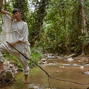 May include: A woman wearing a white, ruffled top and white pants stands in a shallow stream in a lush green forest. She holds a long, thin branch in her hand.