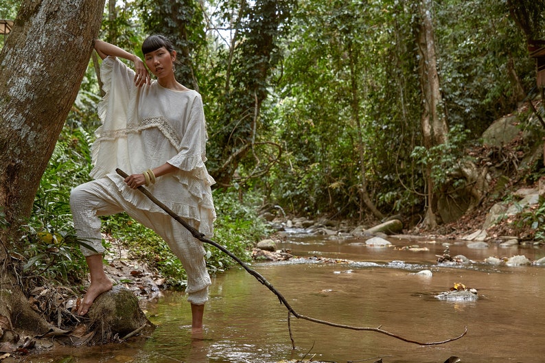 May include: A woman wearing a white, ruffled top and white pants stands in a shallow stream in a lush green forest. She holds a long, thin branch in her hand.