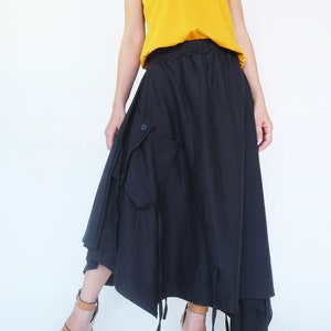 May include: A black maxi skirt with a drawstring waist and a large cargo pocket on the side. The skirt is asymmetrical and has a flowing, relaxed fit. The model is wearing a mustard yellow tank top and brown wedge sandals.