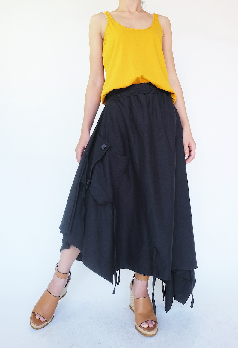 May include: A black maxi skirt with a drawstring waist and a large cargo pocket on the side. The skirt is asymmetrical and has a flowing, relaxed fit. The model is wearing a mustard yellow tank top and brown wedge sandals.