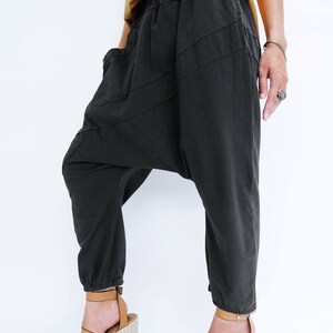 Pu&ograve; includere: Pantaloni neri larghi con un design asimmetrico unico. I pantaloni hanno una gamba larga e una vita elastica a pieghe. La modella indossa i pantaloni con sandali a zeppa marroni.