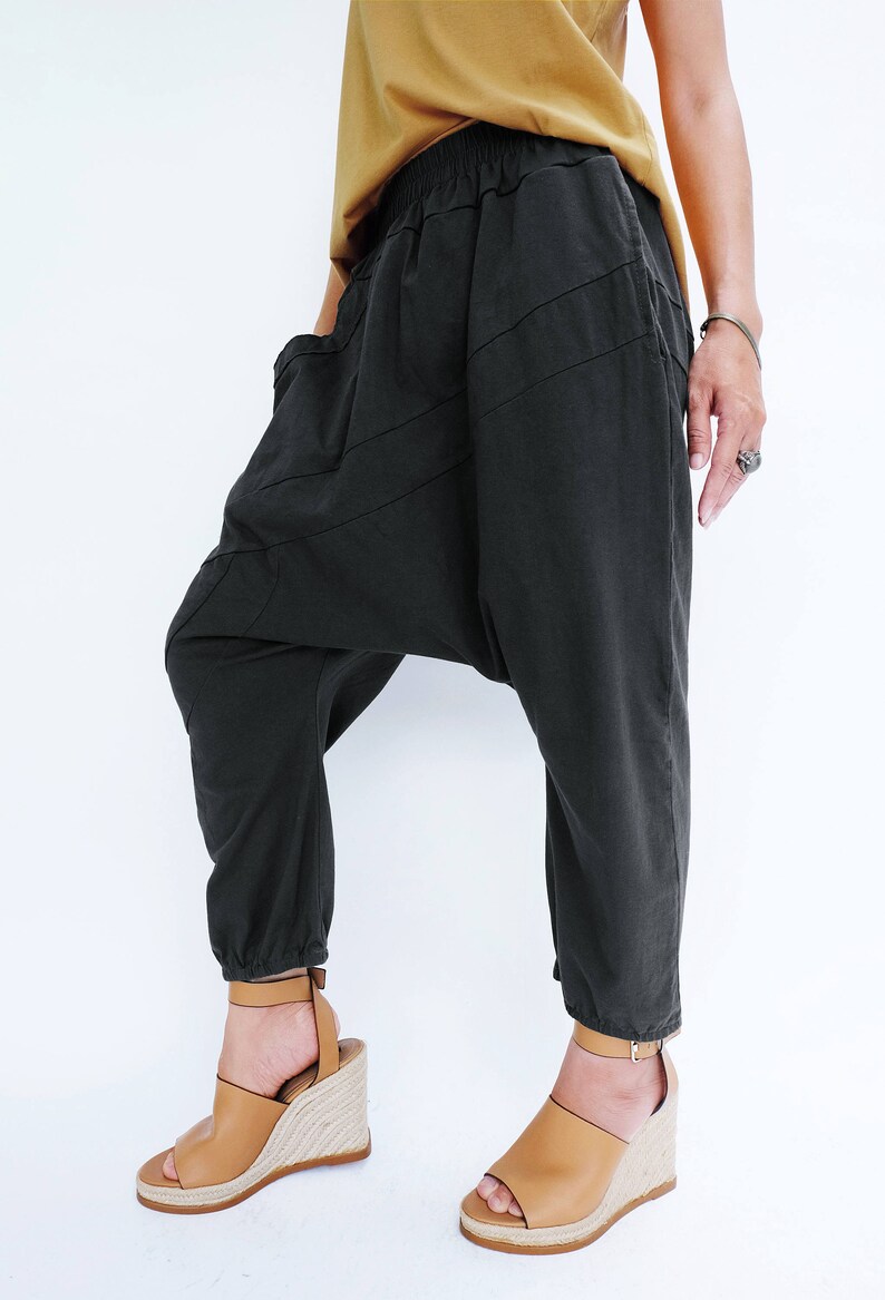 Pu&ograve; includere: Pantaloni neri larghi con un design asimmetrico unico. I pantaloni hanno una gamba larga e una vita elastica a pieghe. La modella indossa i pantaloni con sandali a zeppa marroni.