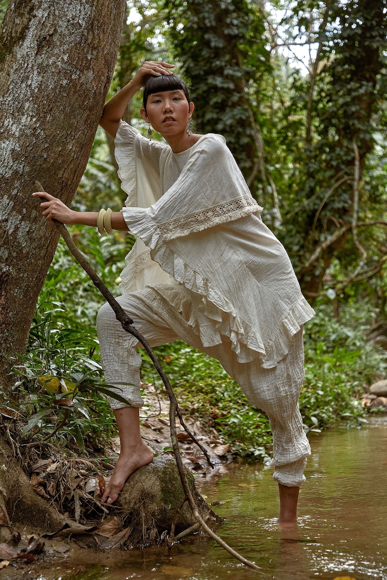 May include: A woman wearing a white, ruffled, crocheted top and white pants stands in a stream in a forest. She is holding a branch with one hand and has her other hand on her head.