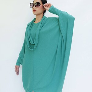 K&ouml;nnte beinhalten: Ein teal-farbenes, lang&auml;rmliges Kleid mit einem Wasserfallausschnitt und einer lockeren Passform. Das Kleid hat einen geschlitzten Saum und wird mit einer gro&szlig;en, quadratischen Sonnenbrille kombiniert. Das Model tr&auml;gt das Kleid vor wei&szlig;em Hintergrund.