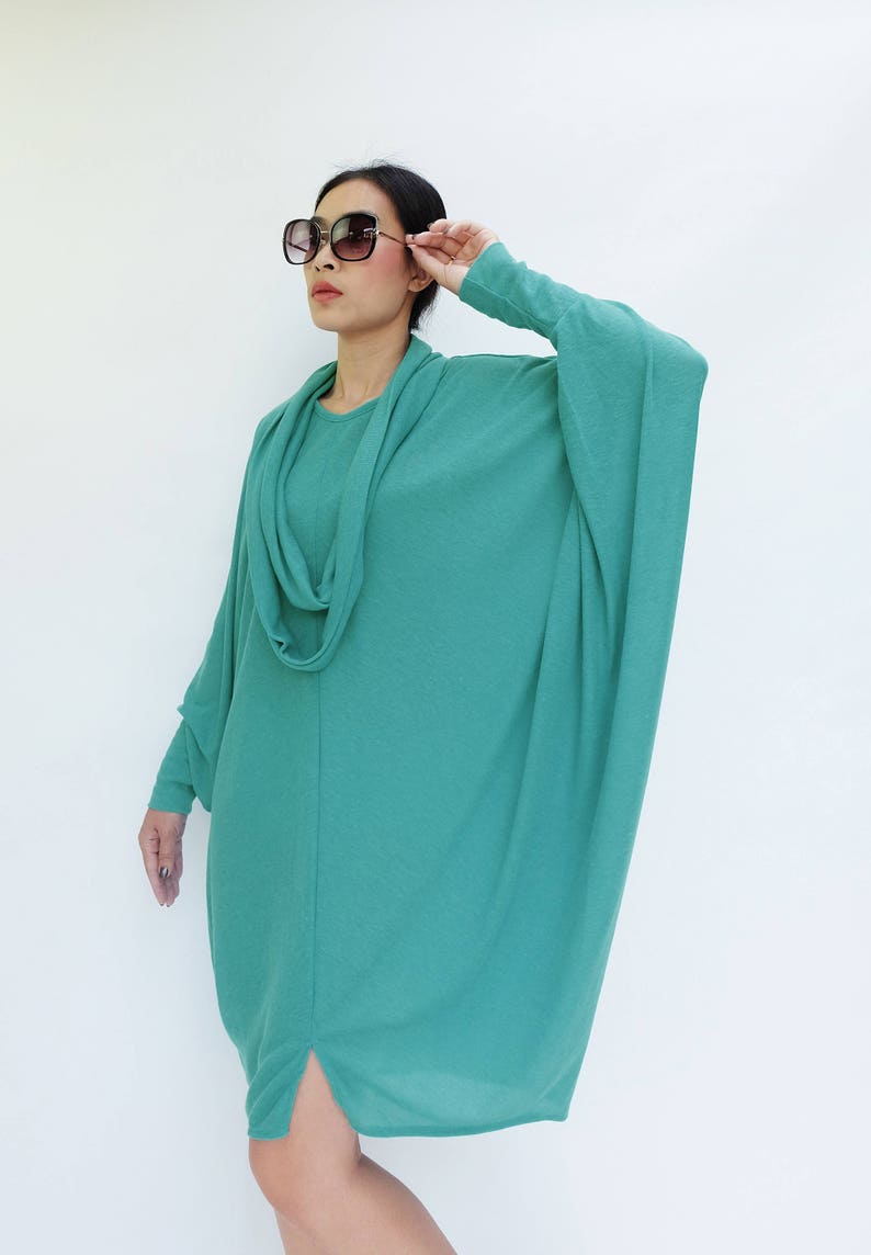 K&ouml;nnte beinhalten: Ein teal-farbenes, lang&auml;rmliges Kleid mit einem Wasserfallausschnitt und einer lockeren Passform. Das Kleid hat einen geschlitzten Saum und wird mit einer gro&szlig;en, quadratischen Sonnenbrille kombiniert. Das Model tr&auml;gt das Kleid vor wei&szlig;em Hintergrund.