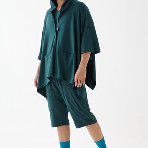May include: A dark green hooded shirt with a long, flowing design and matching shorts. The shirt has a button closure and a pocket on the front. The person is wearing teal socks and white sandals.