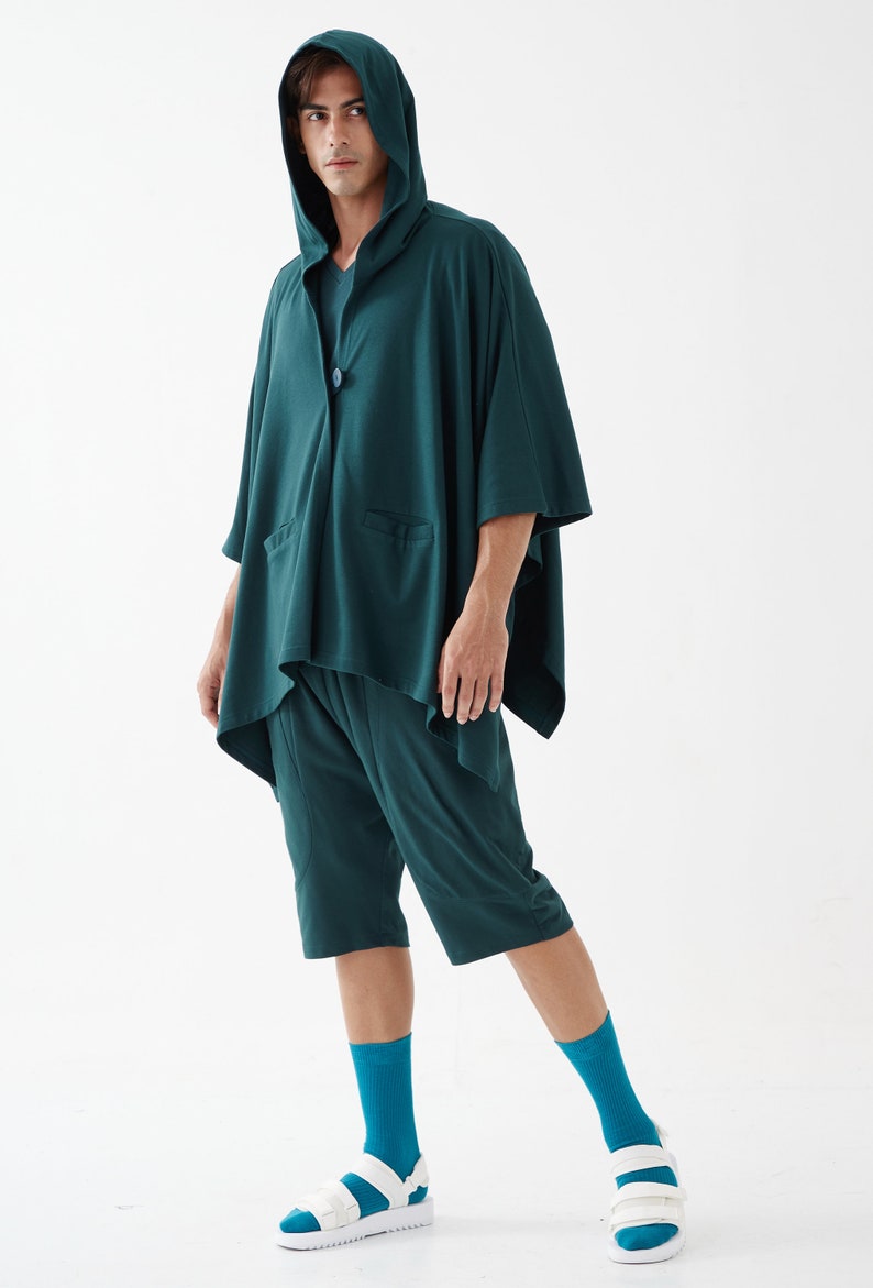May include: A dark green hooded shirt with a long, flowing design and matching shorts. The shirt has a button closure and a pocket on the front. The person is wearing teal socks and white sandals.
