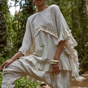 May include: A woman wearing a white, ruffled, crochet top and matching pants stands in a forest setting. She is wearing a pair of large, cream-colored bangles on her wrist.