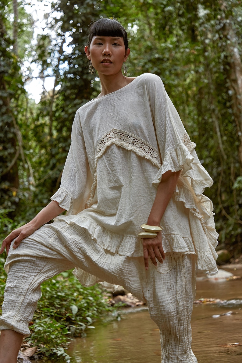 May include: A woman wearing a white, ruffled, crochet top and matching pants stands in a forest setting. She is wearing a pair of large, cream-colored bangles on her wrist.