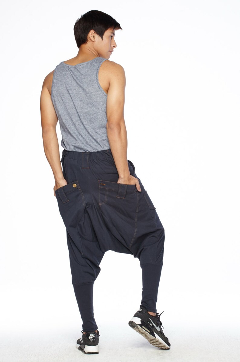 May include: A person wearing a gray tank top and dark blue pants with a unique design. The pants have a loose fit and a gathered look around the knees. The person is wearing black sneakers.