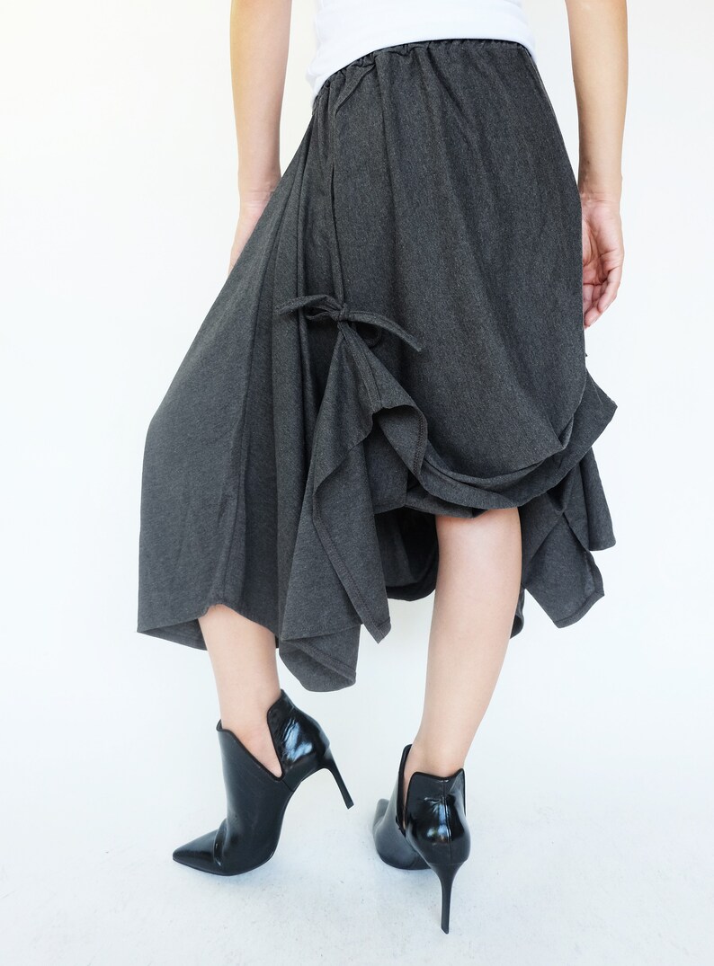 May include: A charcoal gray asymmetrical skirt with a gathered detail at the front. The skirt is made of a soft, drapey fabric and has a relaxed fit. The model is wearing the skirt with black heeled ankle boots.