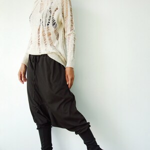 May include: A woman is wearing a white, distressed, long-sleeved sweater and black, loose-fitting pants with a drop crotch.