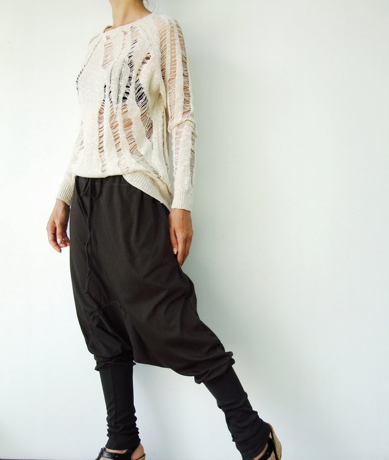 May include: A woman is wearing a white, distressed, long-sleeved sweater and black, loose-fitting pants with a drop crotch.