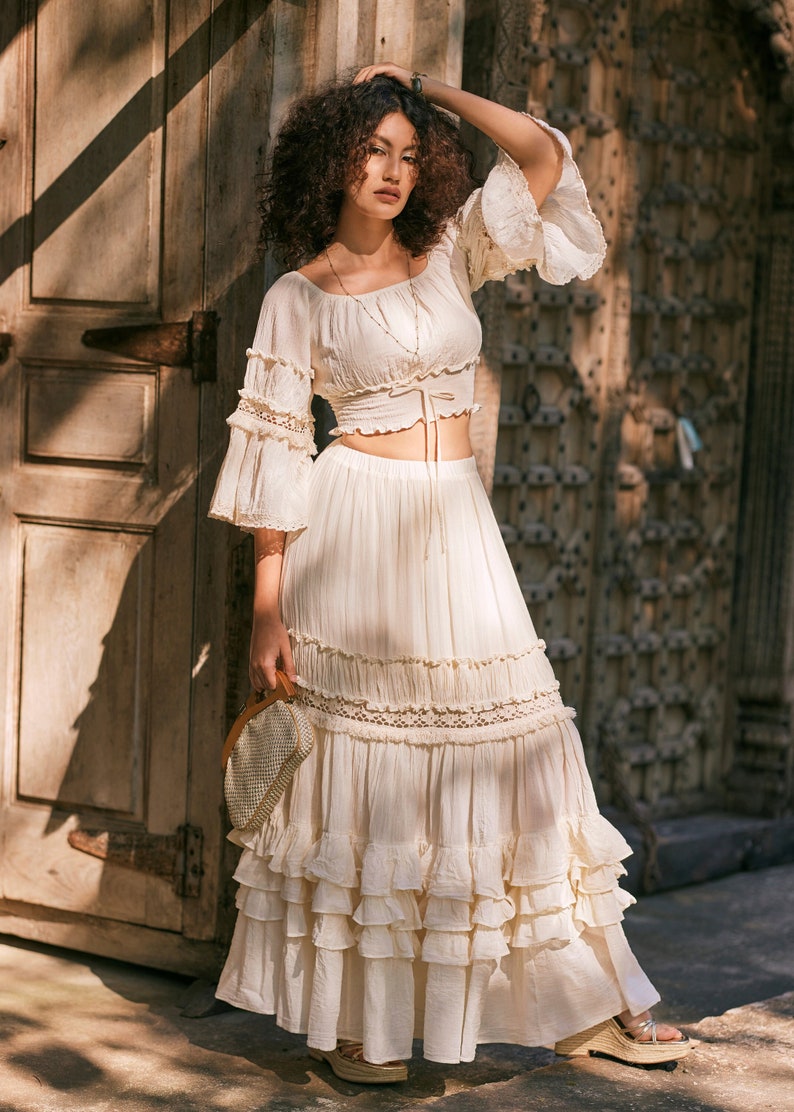 Puede incluir: Un top blanco sin hombros con cuello de volantes y mangas acampanadas, combinado con una falda maxi blanca con m&uacute;ltiples niveles de volantes y encaje. La falda tiene una cintura con cord&oacute;n.