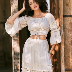 Puede incluir: Un conjunto de top y falda de crochet blanco con escote cuadrado, mangas con volantes y un lazo en la cintura. La falda tiene varias capas y un ribete de encaje.