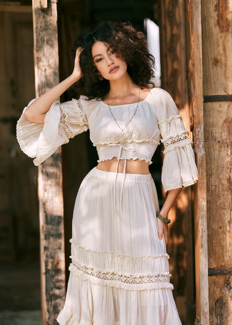Puede incluir: Un conjunto de top y falda de crochet blanco con escote cuadrado, mangas con volantes y un lazo en la cintura. La falda tiene varias capas y un ribete de encaje.