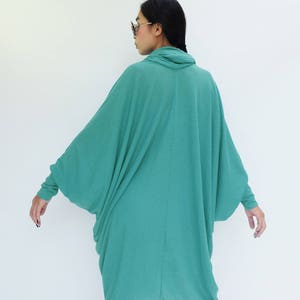 K&ouml;nnte beinhalten: Ein teal-farbenes, lang&auml;rmeliges Kleid mit einem Rollkragen. Das Kleid hat ein lockeres, flie&szlig;endes Design mit weiten &Auml;rmeln und einem drapierten Rock. Das Model tr&auml;gt eine Sonnenbrille. Das Kleid ist aus einem weichen, leichten Stoff gefertigt.