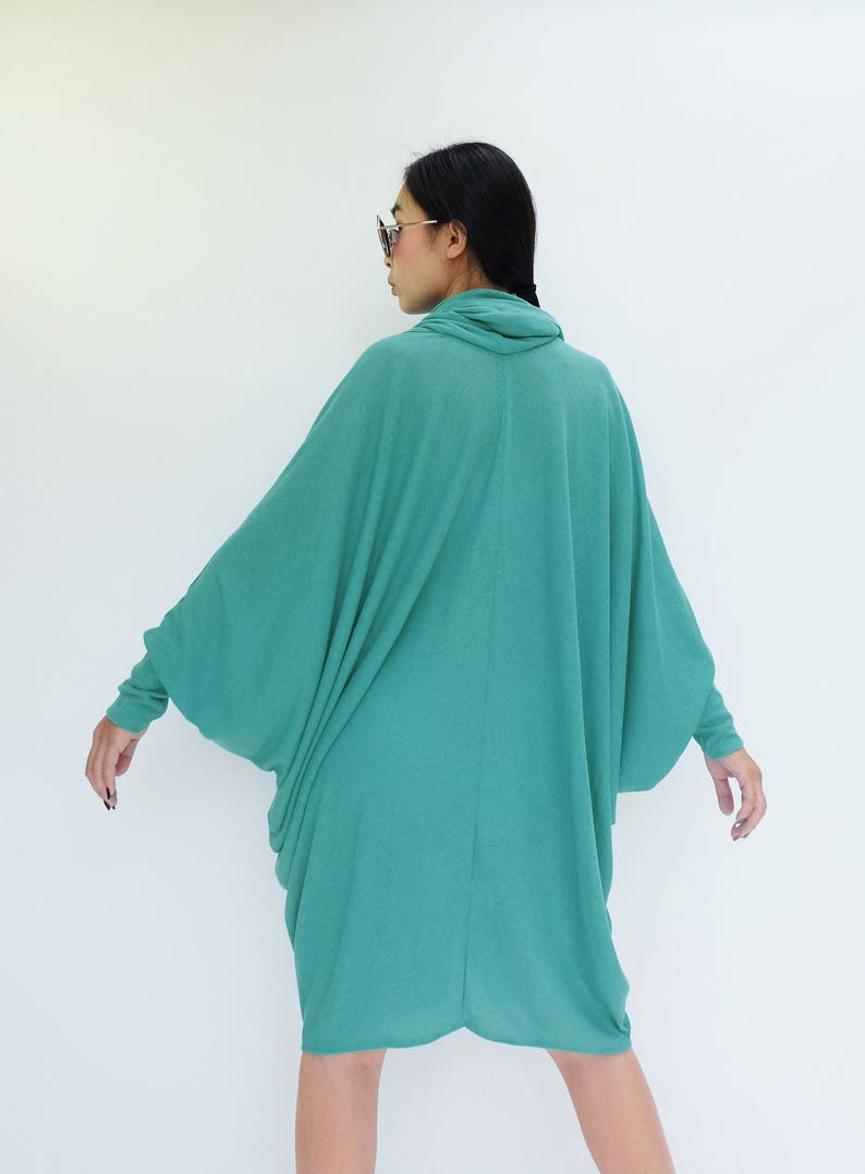 K&ouml;nnte beinhalten: Ein teal-farbenes, lang&auml;rmeliges Kleid mit einem Rollkragen. Das Kleid hat ein lockeres, flie&szlig;endes Design mit weiten &Auml;rmeln und einem drapierten Rock. Das Model tr&auml;gt eine Sonnenbrille. Das Kleid ist aus einem weichen, leichten Stoff gefertigt.