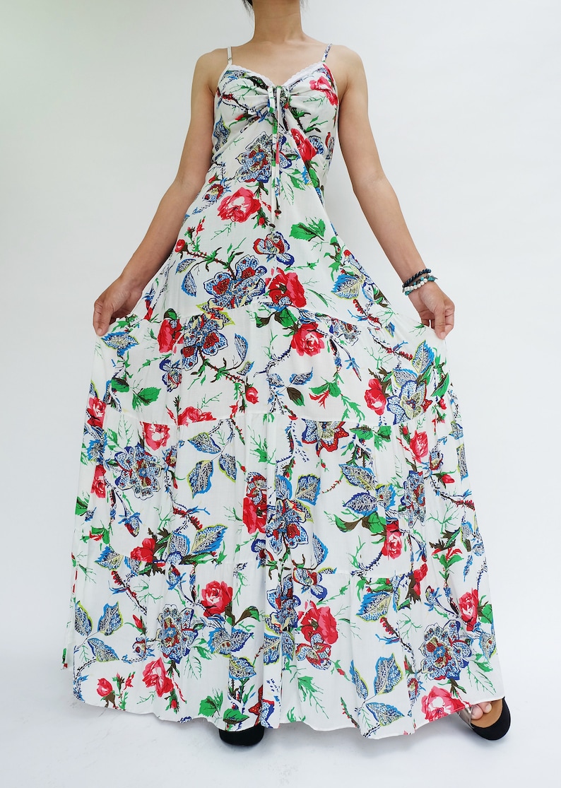 May include: A white maxi dress with a floral pattern featuring red and blue flowers and green leaves. The dress has spaghetti straps and a tiered skirt. The dress is a summer or beach style.