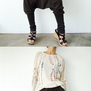 May include: A woman is wearing a white, open-knit sweater and brown, loose-fitting pants with a drawstring waist. She is wearing black sandals with cork soles.