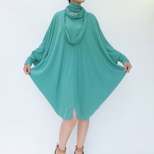 K&ouml;nnte beinhalten: T&uuml;rkisfarbenes Kleid mit langen &Auml;rmeln, Rollkragen und passendem Schal. Das Kleid hat eine lockere, flie&szlig;ende Form und reicht bis zu den Knien. Die Person tr&auml;gt braune Keilsandalen. Der Hintergrund ist schlicht wei&szlig;.