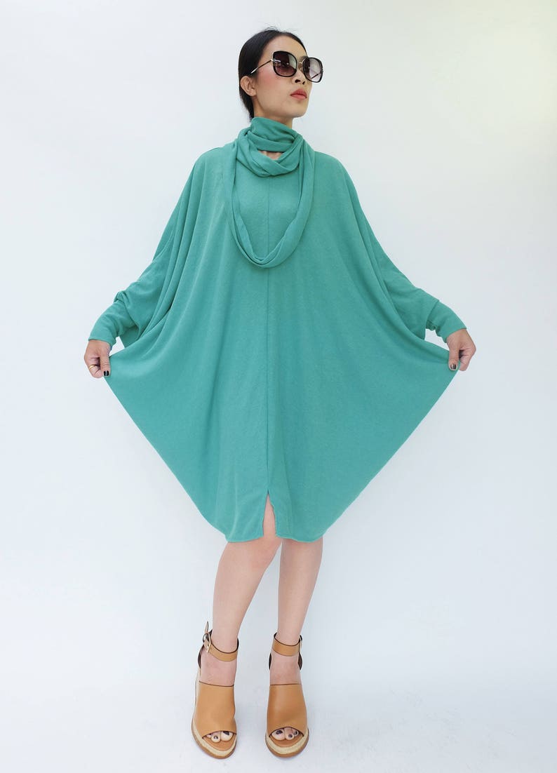 K&ouml;nnte beinhalten: T&uuml;rkisfarbenes Kleid mit langen &Auml;rmeln, Rollkragen und passendem Schal. Das Kleid hat eine lockere, flie&szlig;ende Form und reicht bis zu den Knien. Die Person tr&auml;gt braune Keilsandalen. Der Hintergrund ist schlicht wei&szlig;.